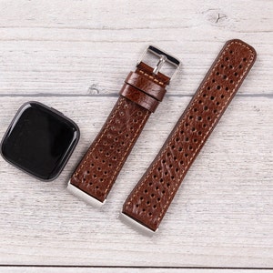 May include: A brown leather watch band with a perforated design and stainless steel hardware. The band is next to a black smartwatch with a square face. The watch band is shown on a light wood grain surface.