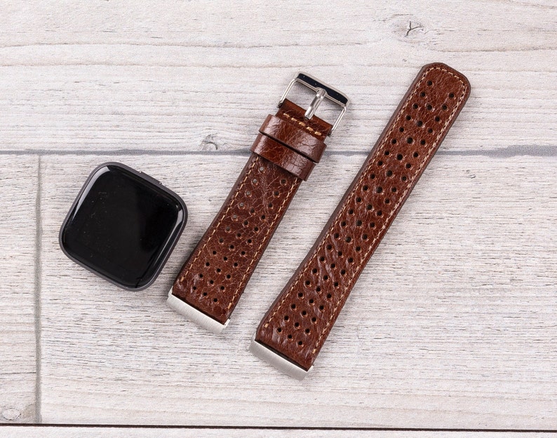 May include: A brown leather watch band with a perforated design and stainless steel hardware. The band is next to a black smartwatch with a square face. The watch band is shown on a light wood grain surface.