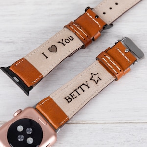 May include: Two brown leather Apple Watch bands with white stitching. One band is engraved with "I love you" and a heart, the other is engraved with "BETTY" and a star.