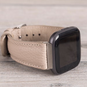 Puede incluir: Una correa de reloj de cuero beige con un reloj inteligente cuadrado negro adjunto. La correa del reloj tiene un cierre de hebilla y detalles de costura.