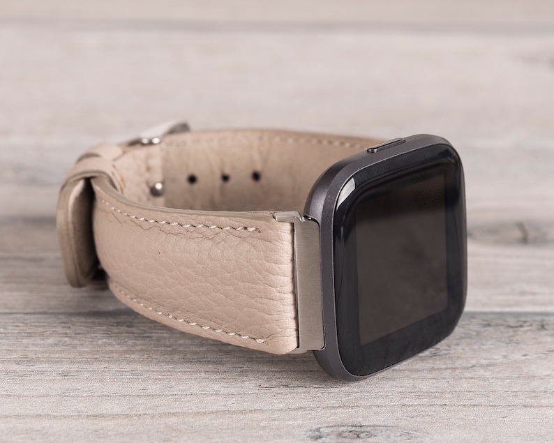 Puede incluir: Una correa de reloj de cuero beige con un reloj inteligente cuadrado negro adjunto. La correa del reloj tiene un cierre de hebilla y detalles de costura.
