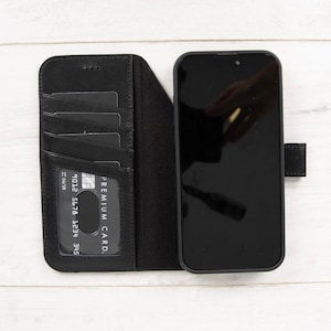 May include: Black leather phone case with card slots. The case is open, revealing several card slots and a black smartphone. The phone screen is black, and the case has a magnetic closure.