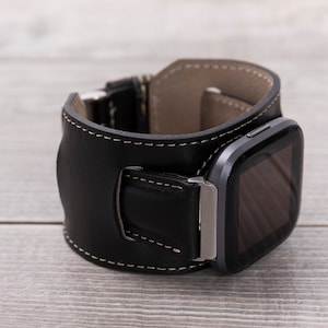 May include: A black leather watch band with white stitching and a silver buckle. The band is designed to fit a square-shaped smartwatch.