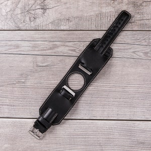 May include: Black leather watch strap with white stitching and a silver buckle. The strap has a hole for a watch face.