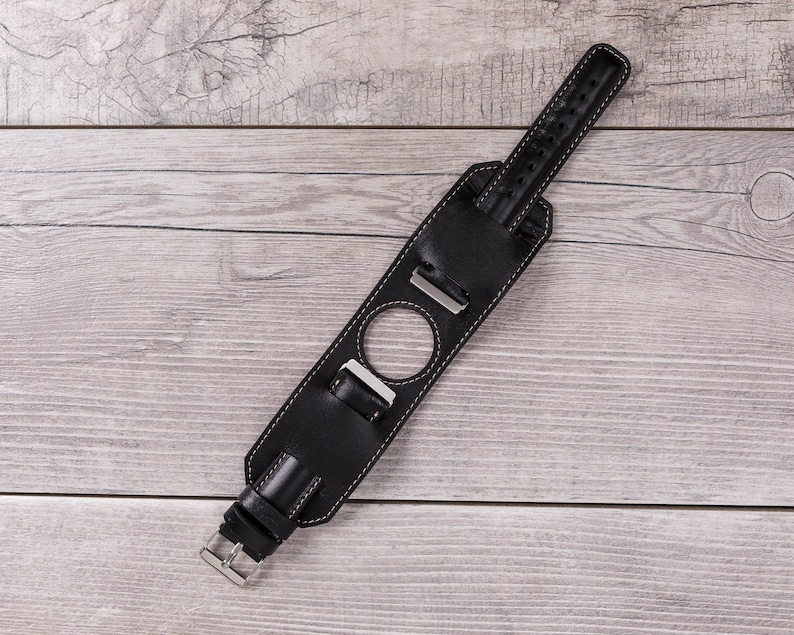 May include: Black leather watch strap with white stitching and a silver buckle. The strap has a hole for a watch face.
