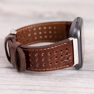 May include: Brown leather watch band with a perforated design. The band is attached to a dark gray smartwatch with a silver clasp. The watch band is made of brown leather with white stitching.