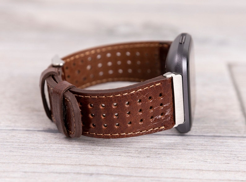 May include: Brown leather watch band with a perforated design. The band is attached to a dark gray smartwatch with a silver clasp. The watch band is made of brown leather with white stitching.