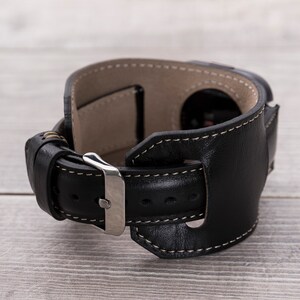 May include: Black leather watch band with a silver buckle and white stitching. The band has a unique design with a leather strap that wraps around the wrist.