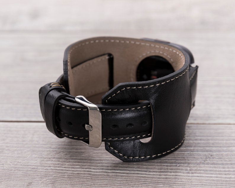 May include: Black leather watch band with a silver buckle and white stitching. The band has a unique design with a leather strap that wraps around the wrist.