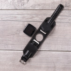 May include: A black leather watch band with a silver buckle and a round cutout for a watch face. The band is lying on a light wood surface.
