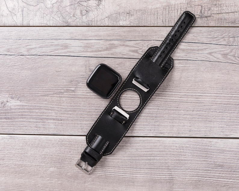May include: A black leather watch band with a silver buckle and a round cutout for a watch face. The band is lying on a light wood surface.