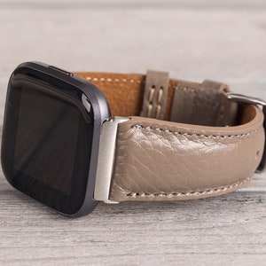 May include: A smartwatch with a square black screen and a taupe leather band. The band is stitched with white thread and has a silver buckle. The watch is resting on a light gray wooden surface.