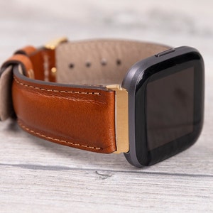 Könnte beinhalten: Ein braunes Lederarmband mit goldener Akzent und eine schwarze Smartwatch mit quadratischem Zifferblatt.