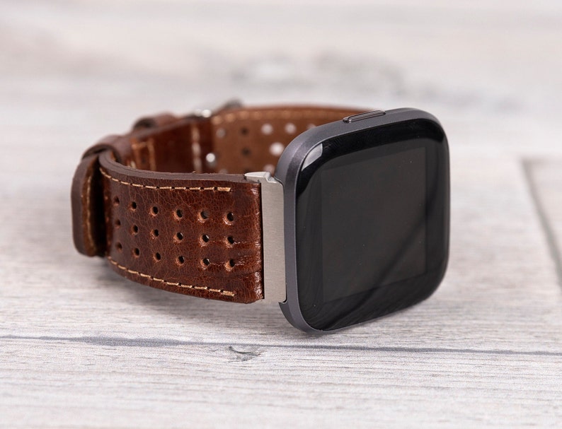 May include: A brown leather watch band with a perforated design is attached to a black smartwatch. The band has a stitched edge and a silver clasp. The watch face is black and rectangular.