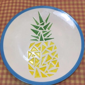 Könnte beinhalten: Ein weißer Keramikteller mit blauem Rand zeigt ein gelbes und grünes Ananas-Design. Die Ananas besteht aus geometrischen Formen.