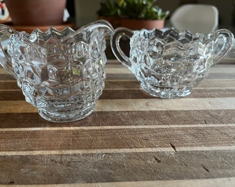 Peut inclure: Deux pots à crème et à sucre en verre transparent avec un motif en diamant. Les pots sont sur une surface en bois.