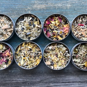 Könnte beinhalten: Acht kleine, runde Metalldosen gefüllt mit verschiedenen getrockneten Kräutern und Blumen. Der Inhalt variiert in den Farben, darunter Grün, Gelb, Rot und Braun. Die Dosen sind auf einer verwitterten, blau-grauen Holzoberfläche angeordnet.