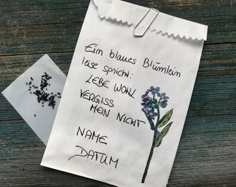 Może przedstawiać: Biały papierowy pakiet z ilustracją kwiatu niezapominajki i tekstem "Ein blaues Blümlein leise sprich: Lebe wohl Vergiss mein nicht NAME DATUM". Obok papierowego pakietu znajduje się mały pakiet z czarnymi nasionami.