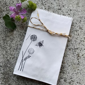 Könnte beinhalten: Eine weiße Papiertüte, gebunden mit gold-weißem Bindfaden, mit einer schwarzen Illustration von Löwenzahnpflanzen und einer Biene. Eine lila Blume und Grünzeug befinden sich im Hintergrund.