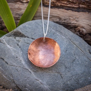 May include: A copper pendant necklace with a round, slightly concave shape hangs on a silver chain. The pendant is resting on a grey rock.