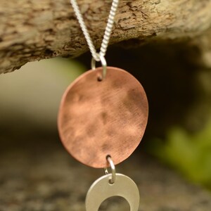 Pu&ograve; includere: Una collana con catena d'argento con un ciondolo a disco color rame e un ciondolo a mezzaluna d'argento. La collana &egrave; esposta su uno sfondo di legno naturale, evidenziando il contrasto tra i metalli e l'ambiente organico.