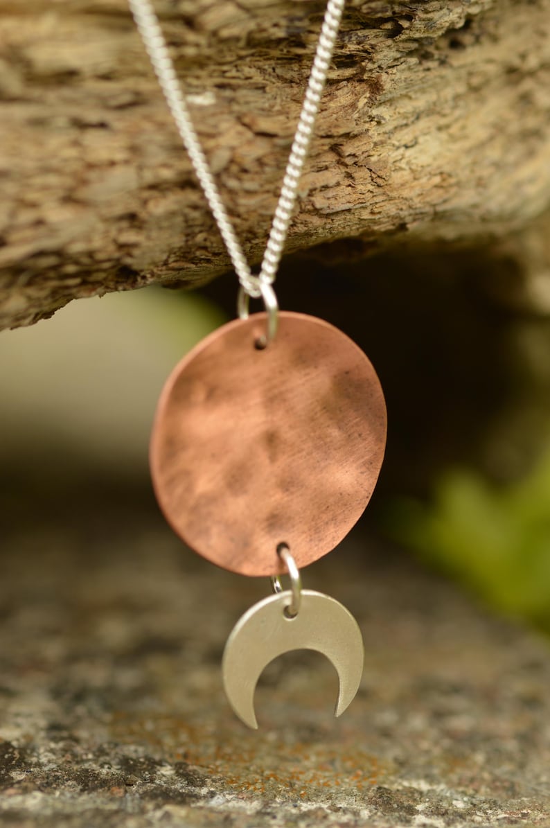 Pu&ograve; includere: Una collana con catena d'argento con un ciondolo a disco color rame e un ciondolo a mezzaluna d'argento. La collana &egrave; esposta su uno sfondo di legno naturale, evidenziando il contrasto tra i metalli e l'ambiente organico.
