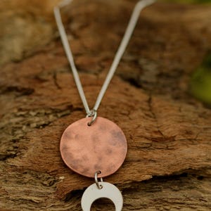 Pu&ograve; includere: Collana con catena d'argento, pendente a disco color rame e ciondolo a mezzaluna argentata. La collana &egrave; esposta su una superficie di legno strutturata, evidenziando il contrasto tra i metalli e lo sfondo naturale.