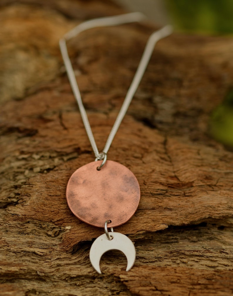 Pu&ograve; includere: Collana con catena d'argento, pendente a disco color rame e ciondolo a mezzaluna argentata. La collana &egrave; esposta su una superficie di legno strutturata, evidenziando il contrasto tra i metalli e lo sfondo naturale.