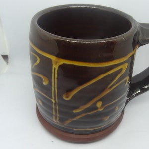 May include: A brown ceramic mug with a yellow glaze design. The mug has a handle and a rounded base.