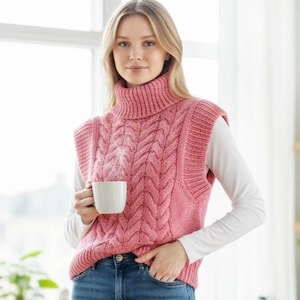 May include: A pink cable-knit turtleneck sweater vest. The vest has a high, ribbed collar and a textured, woven design. The person is wearing a white long-sleeved shirt underneath and blue jeans. A white mug is held in the person's hand.