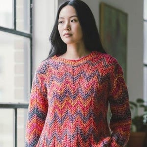 May include: A colorful knit sweater with a chevron pattern in shades of pink, orange, purple, and blue. The sweater has a round neckline and long sleeves. The yarn appears to be a thick, textured wool, perfect for cold weather.