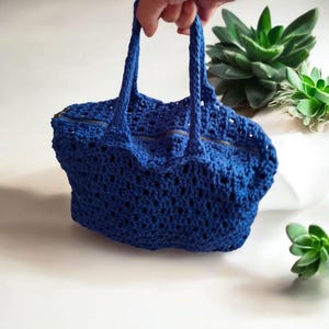 May include: A blue crocheted tote bag with a zipper closure and two handles. The bag is made with a delicate open stitch pattern.