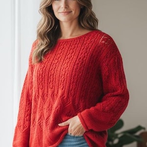 May include: A woman wearing a red knit sweater with a textured pattern and long sleeves. The sweater has a round neckline and is paired with blue jeans. The woman is standing indoors near a window.