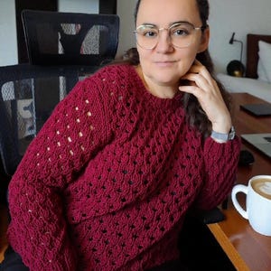 May include: A burgundy-colored, crew-neck sweater with a textured, open-weave design. The sweater has long sleeves and is made of a soft, knitted material. A white mug with a coffee drink is on a wooden desk.