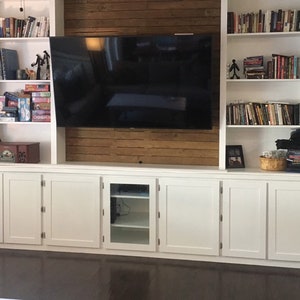 May include: White built-in entertainment center with a flat screen TV, shelves filled with books, and white cabinets with glass doors.