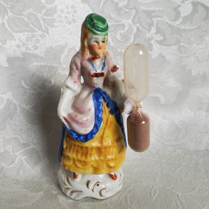 May include: A porcelain figurine of a woman wearing a pink and yellow dress and a green hat, holding an hourglass with brown sand.