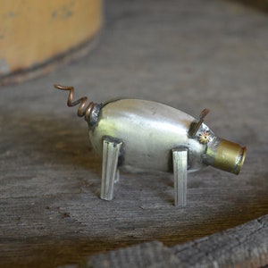 Puede incluir: Una pequeña escultura de cerdo de plata hecha de metal reciclado. El cerdo tiene una cola de cobre rizada, un hocico de bala y una pequeña flor de metal en la cabeza.
