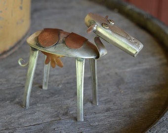 May include: A whimsical metal sculpture of a cow made from recycled spoons and other metal scraps. The cow is brown and silver with a curly tail and a friendly face.