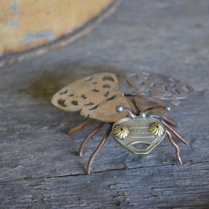 May include: A whimsical insect sculpture made from repurposed metal, featuring a key for the body and two ornate metal pieces for wings. The insect has copper wire legs and gold-colored eyes.