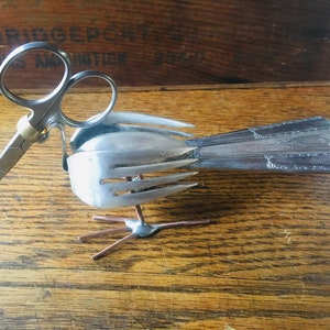 Puede incluir: Una escultura de pájaro caprichosa hecha de cubiertos reciclados. El cuerpo está formado por tenedores, la cabeza por unas tijeras y la cola por cucharas. Alambre de cobre forma las patas, creando una pieza única y artística.