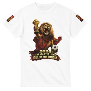 May include: White t-shirt featuring a roaring lion holding a trophy and a soccer ball. The lion is wearing a red cape. Text on the shirt reads "2026: THE YEAR THE LIONS RULED THE JUNGLE."