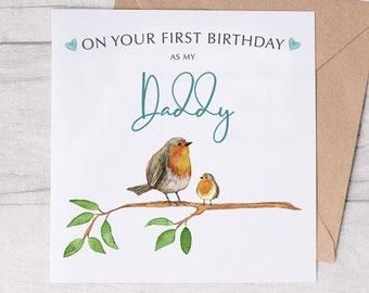 Op de afbeelding: Een witte kaart met een blauw en groen hart aan elke kant van de tekst "On your first birthday as my Daddy". De kaart heeft een aquarel-illustratie van twee roodborstjes die op een tak zitten met groene bladeren.