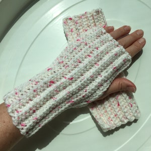 May include: White crocheted fingerless gloves with pink flecks. The gloves are shown on a hand, with the fingers exposed.