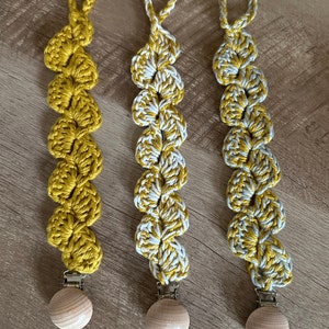 May include: Three crocheted pacifier clips with wooden beads. The clips are made with yellow and white yarn and feature a heart-shaped design.
