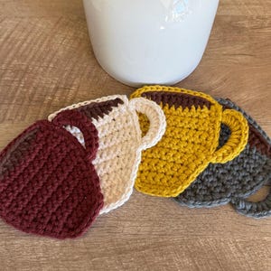 Könnte beinhalten: Eine Sammlung handgefertigter Häkeluntersetzer in verschiedenen Farben, darunter Burgunderrot, Creme, Gelb und Grau. Jeder Untersetzer ist wie eine Kaffeetasse mit Henkel geformt. Die Untersetzer sind auf einer Holzoberfläche neben einer weißen Keramikvase angeordnet.