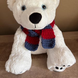 May include: A plush white polar bear stuffed animal with soft fur, round ears, and light blue eyes. The bear has a black nose and is wearing a hand-crocheted scarf featuring horizontal stripes in alternating dark red and navy blue yarn colors.
