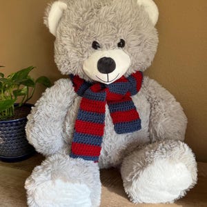 May include: A large, plush gray teddy bear with white accents on the paws and ears. The bear wears a crocheted scarf with red and navy blue stripes. The bear has a black nose and a smiling expression.