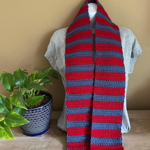 May include: A handmade crochet scarf with alternating red and blue horizontal stripes. The scarf is displayed on a mannequin, showcasing its length and pattern. The scarf is made of soft yarn, perfect for cold weather.