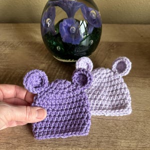 May include: Two handmade crochet mouse hats in shades of purple. One hat is held by a hand. The hats are square with rounded ears. The background includes a glass paperweight with purple floral designs.