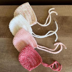 May include: Four handmade crochet baby bonnets in shades of pink and white, with tie closures. The bonnets are arranged on a wooden surface, showcasing the texture and colour variations of the yarn. A charming and delicate baby accessory.